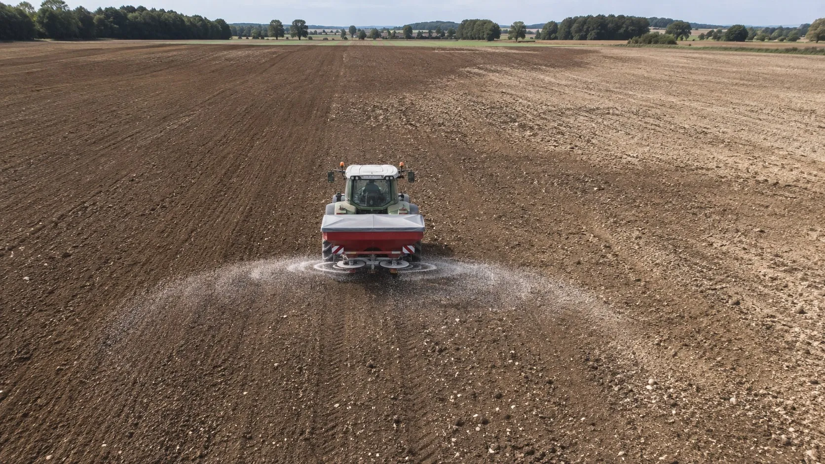 Vue aérienne d’un tracteur et de son épandeur appliquant de l’engrais de fond sur une parcelle en sol nu.
